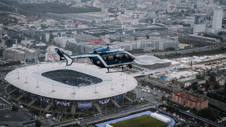 Vue prise du ciel d'un hélicoptère gendarmerie bleu en vol au-dessus du stade de France entouré de bâtiments et d'un terrain de sport.