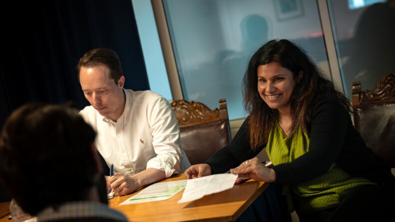 Agrandir l'image Une femme et un homme derrière une table tenant des feuilles; au premier-plan, flou, l'arriere de la la tête d'un homme