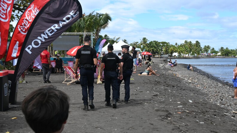 Trois gendarmes mobiles de dos marchant sur une plage de sable noir. à droite la mer. Sur la plage, des personnes assises ou allongées. A gauche des drapeaux publicitaires de Coca COla et Powerade