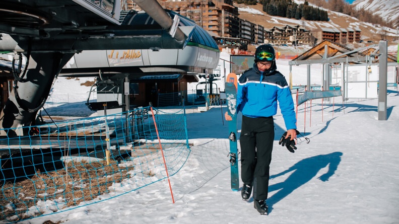 une femme gendarme tenant deux skis de la main droite avance sur la neige, en fond une station de ski