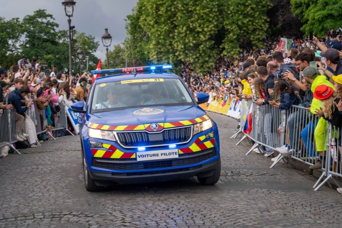 Agrandir l'image Une voiture de la gendarmerie roule dans les rues de Paris, sur une voie pavée, le logn de laquelle est massée une foule imortante, contenue derrière des barrières