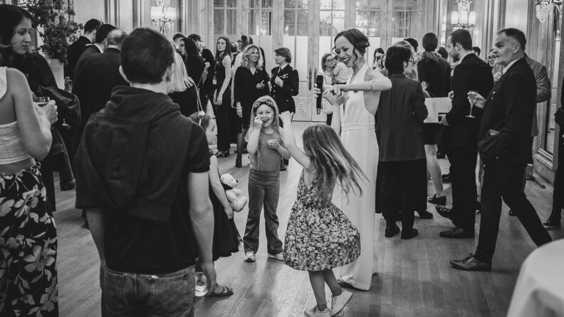 Scèe de fête avec des enfants dansants et adultes conversant, un verre à la main. Au milieu, une femme vêtue d'une combinaison blanche danse avec les enfants