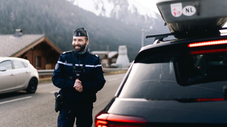 Agrandir l'image Image d'un gendarme souriant, debout près d'une voiture noire, sur le toit duquel est accrochée une galerie. Le militaire vient de procéder à un contrôle du véhicule et du conducteur.