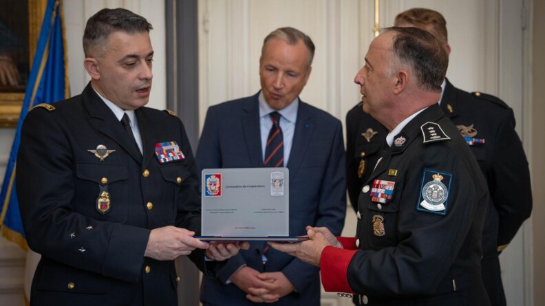 Deux militaires s'échangent un objet gris portant la mention "convention de coopération". Au fond deux hommes en costard