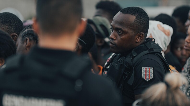 Photo de profil du gendarme Ali, en tenue d'intervention noire, au milieu d'une foule indistincte. Au premier plan, un gendarme de dos, avec la mention GENDARMERIE sur le dos de sa tenue.