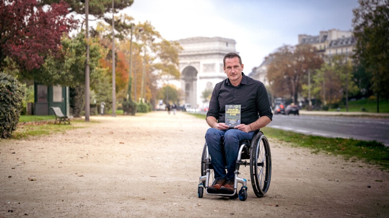 Photo de Franck Robin, prise sur le trottoir, avenue Foch, avec l'Arc de Triomphe en arrière plan. L'ancien militaire, en tenue civile, est en fauteuil roulant. Il tient dans ses mains l'ouvrage dont il est l'auteur.