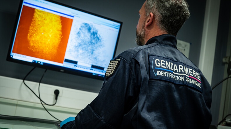 Agrandir l'image Un gendarme de dos, en bleu marine, portant la mention "gendarmerie, identification criminelle", observe un écran d'ordinateur accroché au mur, exposant une empreinte digitale orange et une bleue