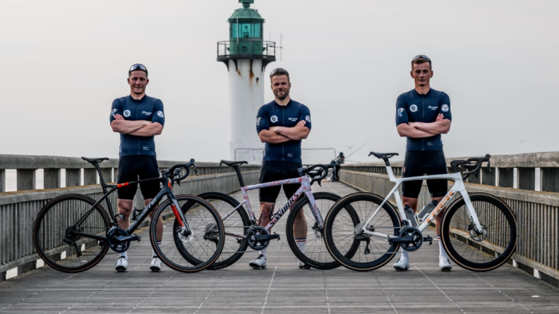 Agrandir l'image Trois hommes en tenue de sport (haut bleu marine et short noir), sur un pont au bot duquel se trouve un phare blanc et vert, debout derrière leurs vélos