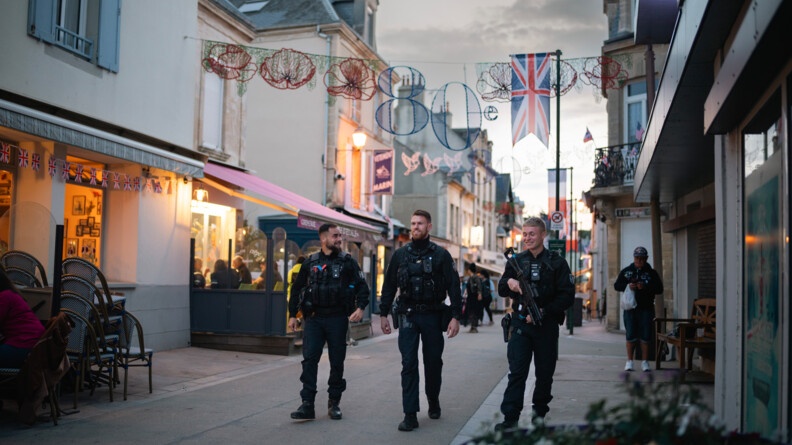 Trois gendarmes marchent dans la rue. Autour d'eux, flottent des drapeaux anglais.