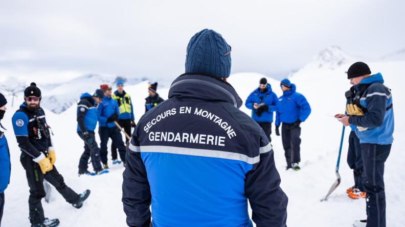 Agrandir l'image Sur un sol enneigné, un groupe d'hommes vêtus d'une tenue de ski de couleur bleue, se tiennent debout en cercle. Derrière eux se dressent les sommets enneigés. Au dos de l'anorak de l'un d'eux, qui se tient au premier plan de la photo, on lit : Secours en montagne Gendarmerie