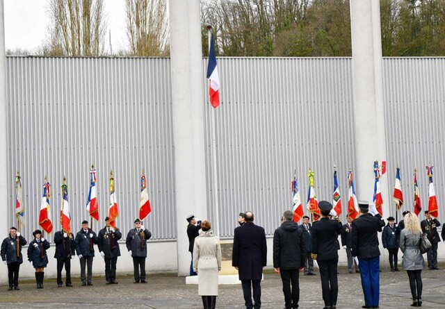 Six personnes, autorités civiles et militaires au salut, vues de dos, face au drapeau en train d'être hissé. De part et d'autre du mât des couleurs, une demi-douzaine de porte-drapeaux.