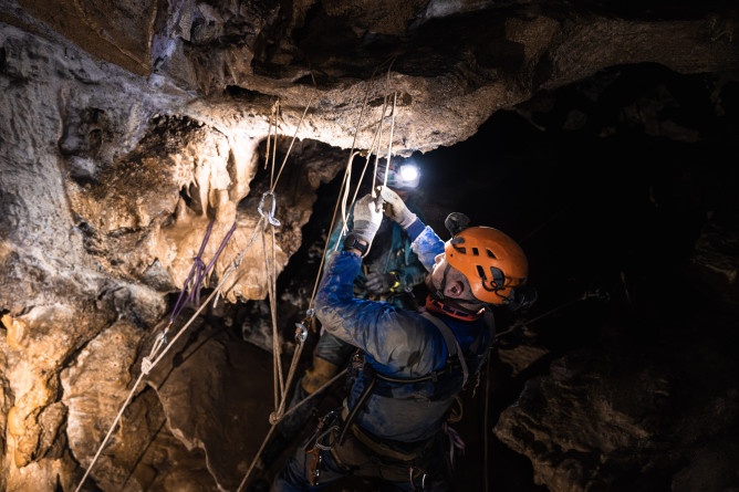 Agrandir l'image Photo montrant un gendarme spéléologue en combinaison et casque équipé d’une lampe frontale, en train d'installer une corde sur une paroi rocheuse à l’intérieur d’une grotte.