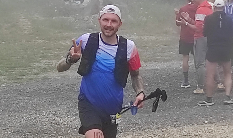 un coureur v&ecirc;tu d'un short noir et d'un maillot bleu blanc rouge, porte des b&acirc;tons de marche dans une main et fait le V de la victoire de l'autre.