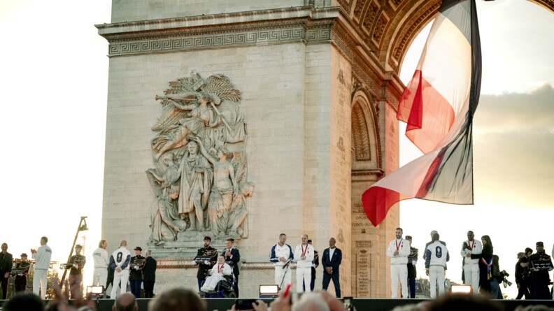 Les athl&egrave;tes des JOP 2024 sur une sc&egrave;ne au pied de l'Arc de Triomphe. Dans le fond des gendarmes en vareuse portent des coussins de d&eacute;corations. Sous l'arche de l'Arc de Triomphe, le drapeau fran&ccedil;ais flotte dans le coucher du soleil.