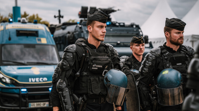 Trois gendarmes mobiles en tenue de maintien de l'ordre, calot sur la tête, patientent devant un véhicule de transport de troupes et un Centaure