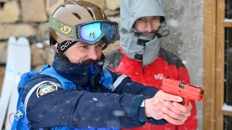 un gendarme coiffé d'un casque de ski pointe un pstolet rouge d'entraînement. en arrière-plan à droite, un homme en rouge