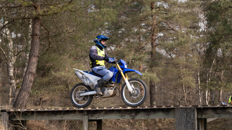 Un motard de la gendarmerie sur une moto cross bleue passe sur un pont de bois dans une forêt de résineux