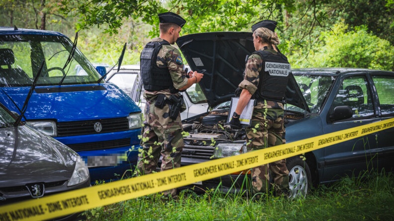 Agrandir l'image Dans une forêt, deux gendarmes sont debout devant une caracasse de voiture sur laquelle il tente de récuperer des élements. En premier plan, se trouve le bandeau jaune de sécurité où il est noté "Gendarmerie nationale - Zone interdite"