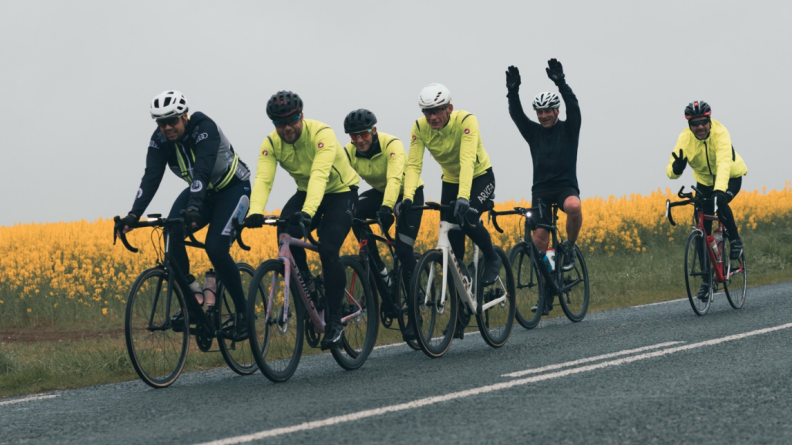 Six cyclistes, le premier en noir, les trois suivant en bas noir et haut jaune fluo, le cinquième en noir, qui pédale et levant les bras, et le sixième en bas noir et haut jaune fluo, faisant un signe "V" de sa main droite. Ils roulent une route goudronnée, le long d'un champ de colza en fleur