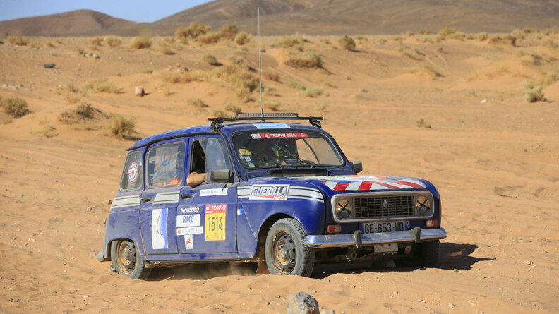 Une Renaud 4L bleu marine, couverte d'enseignes diverses, postionn&eacute;es sur le flanc et le sur capot, roule en plein d&eacute;sert. L'avant-bras du passager d&eacute;passe de la fen&ecirc;tre ouverte.