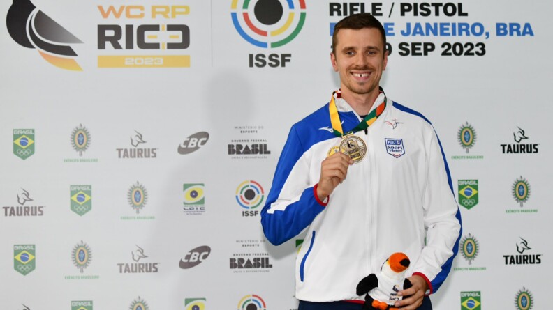 Sur un mur regroupant les sponsors de la coupe du monde de tir à Rio, à droit, un homme en survêtement, souriant, tient s médaille d'or de la main droite et autour du cou, et un toucan en peluche de la main gauche