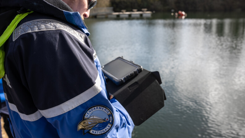 Agrandir l'image Un gendarme au bord d'un plan d'eau regarde un écran dans ses mains