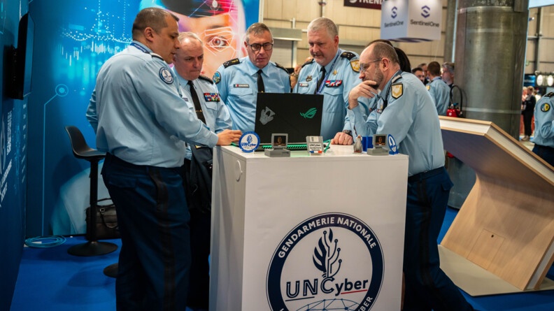 Agrandir l'image Cinq gendarmes, debout autour d'un comptoir, sur lequel est inscrit "Gendarmerie nationale - Unité nationale cyber", regardent l'écran de l'ordinateur posé devant eux. La scène se déroule sur un stand lors d'un salon.