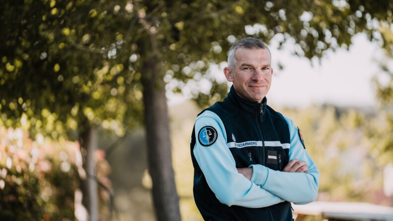 Agrandir l'image Un gendarme un tenue pose face à l'objectif, souriant, bras croisés devant lui, devant un arbre.