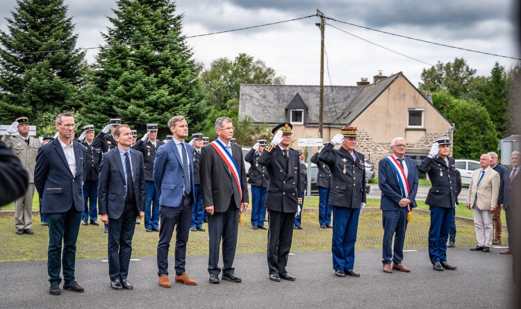 Huit personnes, soit trois &eacute;lus, trois militaires (effectuant un salut) et trois civils, se tiennent debout c&ocirc;te &agrave; c&ocirc;te, dans un village. Autour d'eux sont r&eacute;unis d'autres militaires ainsi que des civils.