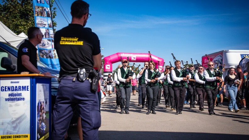 A gauche, le stand de recrutement de la gendarmerie avec deux gendarmes en polo noir siglé en jaune "devenir gendarme". Ils regardent un bagad, dont les musiciens sont vêtus d'une chemise blanche, gilet vert et pantalon noir, et jouent leur instrument en marchant. En arrive plan le point de départ de la course
