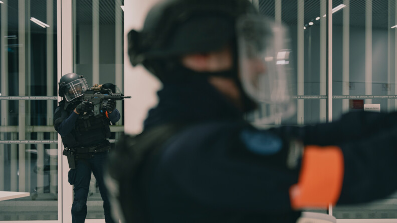 Deux militaires du PSIG Sabre, un de profil au premier plan, pointant une arme de poing, un second en arrière plan armé d'un fusil mitrailleur.