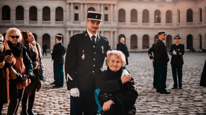 Un gendarme avec une vieille femme sur une chaise avec une b&eacute;quille, s'treignent en regardant l'objectif, dans la cour pav&eacute;e des invalides
