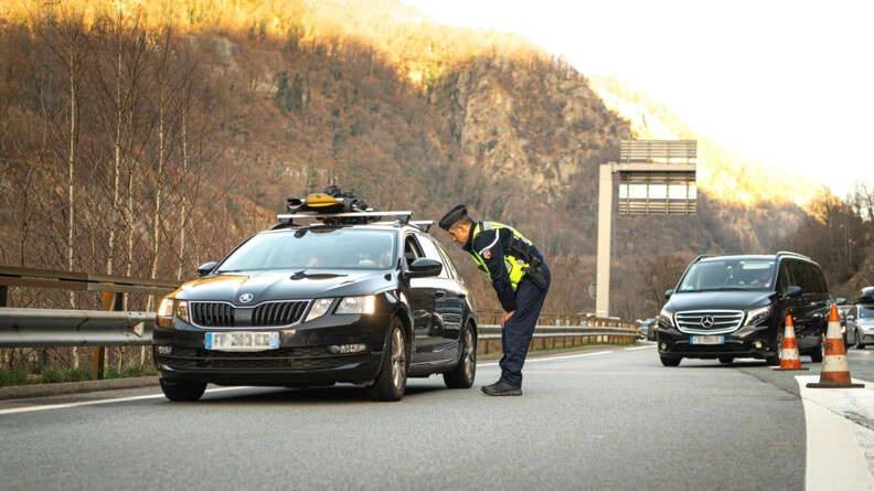 Un gendarme avec une chasuble jaune conseille un automobiliste sur la route. On peut voir des skis sur le toit du véhicule et de nombreuses voitures en arrière plan.