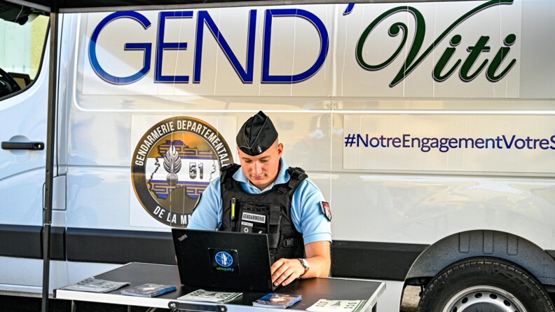 Sous une tonnelle assis à une table noire, derrière un ordinateur portable "ubiquity" et des prospectus , un gendarme assis tape sur l'ordinateur. Derrière lui, une camionnette blanche avec l'inscription Gend'viti
