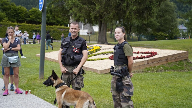 Agrandir l'image Un binôme de maitre de chien gendarmerie, un homme tenant en laisse le malinois et à droite une jeune femme. Ils sont vêtus d'un t-shirt noir et d'un bas de treillis, devant un parterre de fleur au symbole des Jeux olympiques
