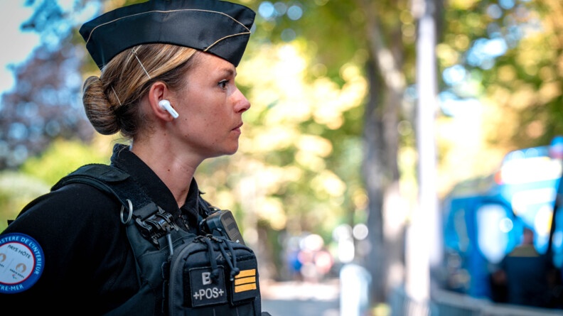 Une jeune femme blonde, dont les cheveux sont coiffés en chignon, vêtue d'un uniforme noir de la gendarmerie, se tient debout, de biais, regardant droit devant elle.