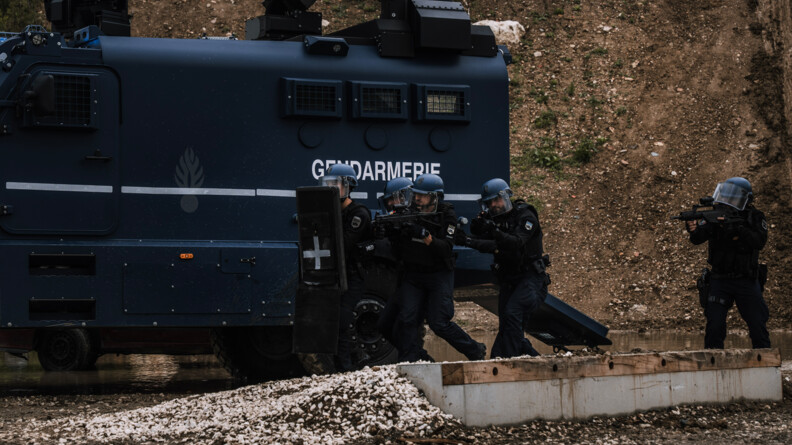 Cinq gendarmes mobiles en colonne d'assaut protégés par un bouclier et équipés d'armes longues remontent le long du Centaure