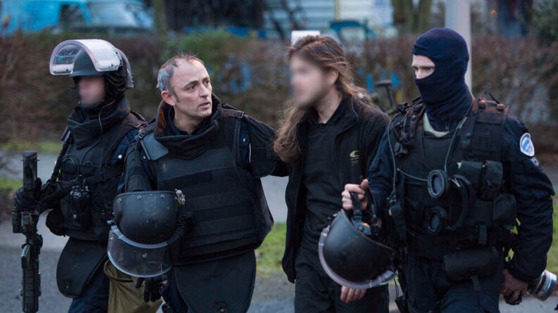 Agrandir l'image Le commandant du GIGN, casque à la main, est aux côtés d'un homme vêtu de noir, cheveux longs, visage flouté. Les deux hommes sont encadrés par deux équipiers du GIGN ; celui à gauche est casqué, cagoulé et tient son fusil à la main ; celui de droite a son casque à la main et porte une cagoule.