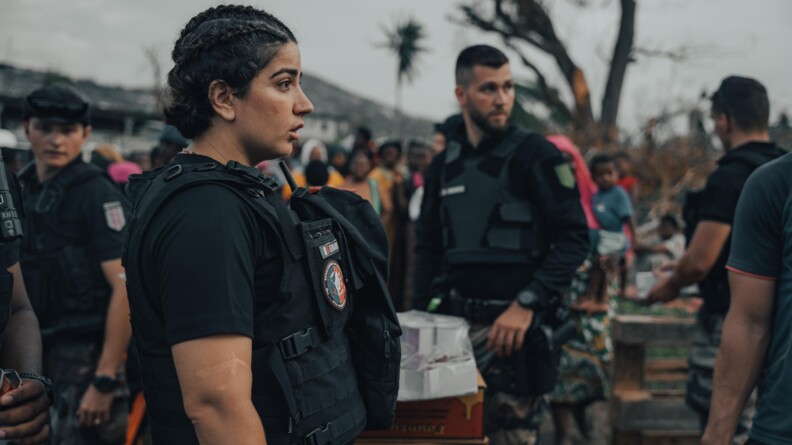 Une gendarme au t-shirt noir et gilet pare balels noire au milieu de gendarmes et de la population en distribution de vivres
