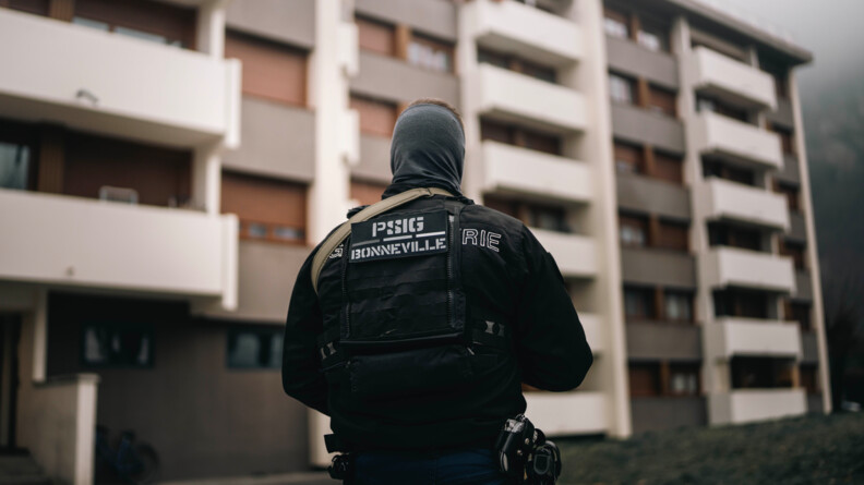 Photo d'un gendarme, debout, de dos, dont le vêtement noir porte l'inscription dans le dos "PSIG Bonneville". Le militaire se tient face à un immeuble.