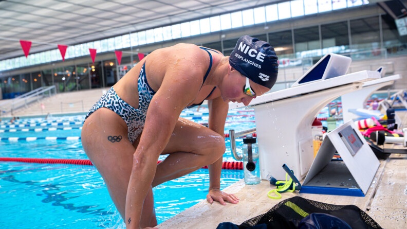 Une nageuse sort d'un bassin olympique, en remontant sur le bord de la piscine.Elle porte un maillot bleu et blanc, un bonnet bleu marine et des lunettes de nage, ainsi qu'un tatouage sur le haut de la cuisse représentant le symbole des Jeux olympiques