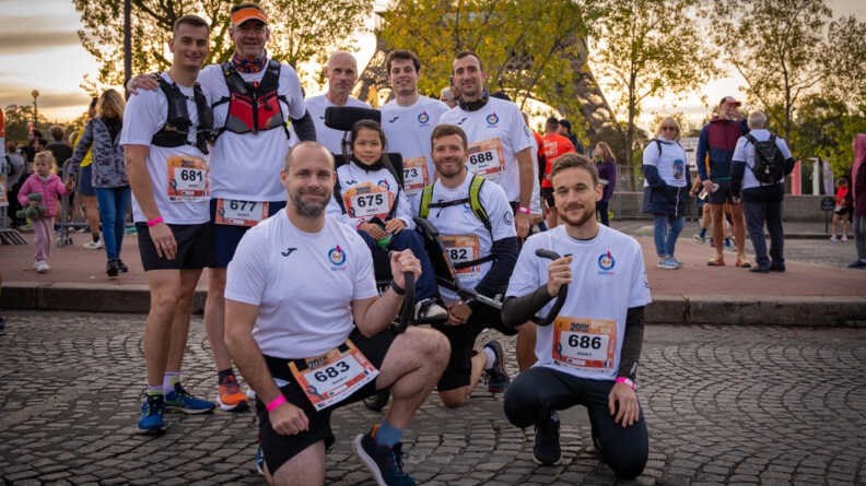 Des coureurs, vêtus d'un T-shirt blanc avec un numéro devant, posent sur une avenue pavée, autour du fauteuil roulant d'une jeune fille d'orgine asiatique. On aperçoit la Tour Eiffel en arrière plan.