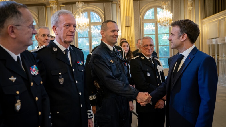 A droite le président de la République, de profil, en costume bleu, souriant, serre la main du général Réty, patron du GIGN, en combinaison GIGN bleue. A la gauche du général, le directeur de la police nationale et le directeur général de la gendarmerie nationale en vareuse..