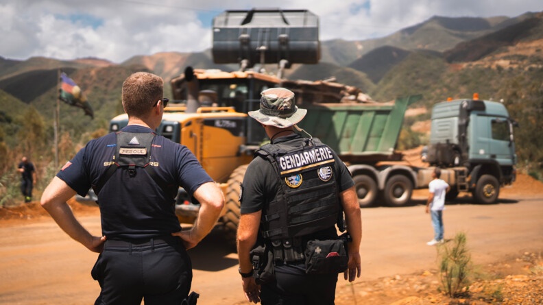 Un agent des FORMISC et un gendarme mobile, vus de dos, observent l'opération de déblaiement de la RT3. Devant eux, un camion-benne et un camion-grue sont à l'oeuvre.