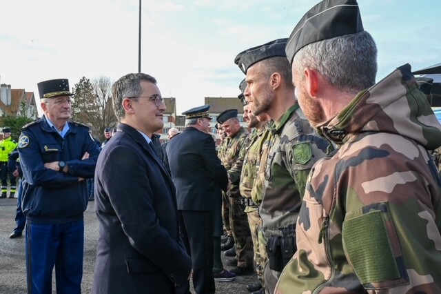 Gérald Darmanin se tient debout, de biais, face à des militaires en tenue kakie. Il regarde l'un d'eux dans les yeux. Derrière le ministre se tient le directeur général de la gendarmerie nationale, en tenue bleue marine, portant un képi et tenant ses bras croisés contre sa poitrine.