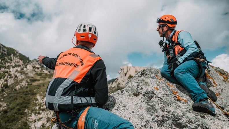 Deux gendarmes sur un rocher, portant une chasuble "secours en montagne", regardant vers les hauteurs