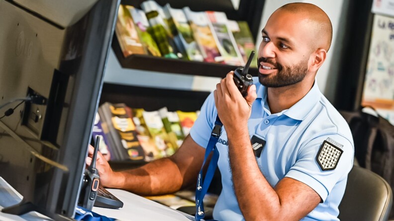 Assis derrière un bureau , souriant, un gendarme tient un talkie-walkie de sa main gauche. Devant lui, un écran d'ordinateur et en arrière-plan un étagère avec des prospectus