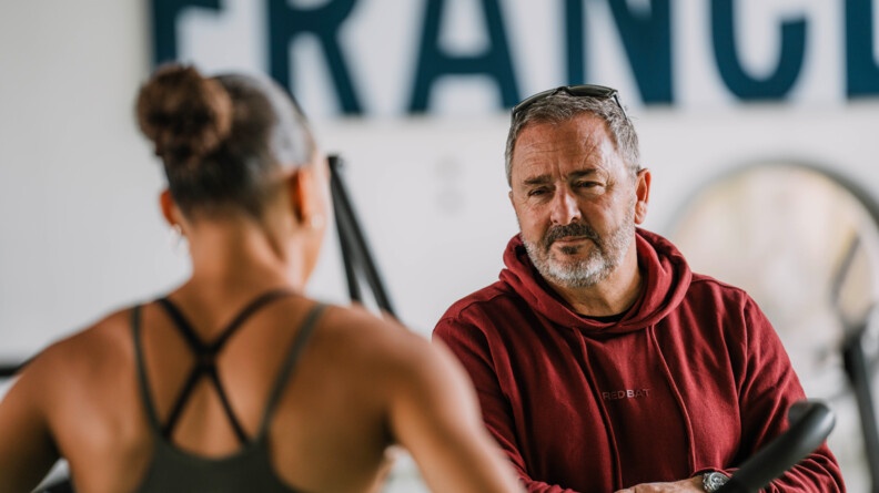Une athlète se tient debout, de dos. Elle fait face à un homme (son entraîneur) qui la regarde. L'homme porte un sweat-shirt à capuche de couleur bordeaux. Sur le mur, derrière eux, on devine écrit en gros, en lettres capitales de couleur bleue marine, le mot FRANCE.