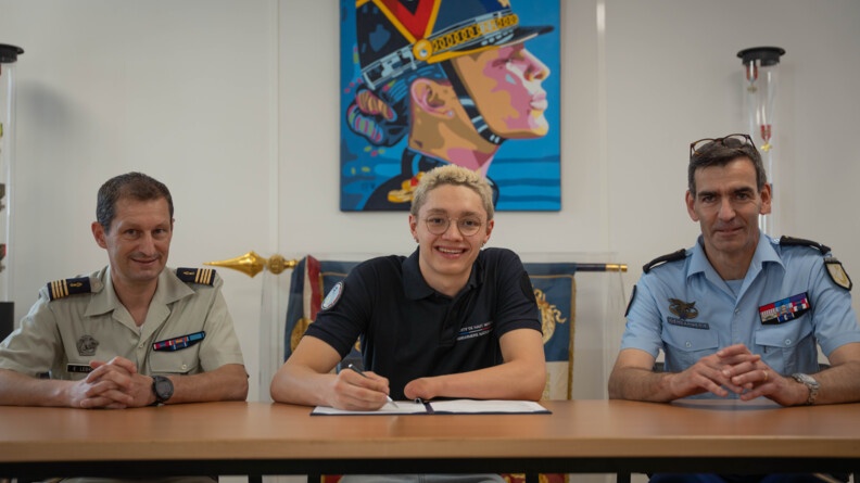 Assis derrière une table, un para-sportif de la haut-niveau de la gendarmerie (SHND-G) souriant et s'apprêtant à signer son contrat de SHND. Sa main gauche est atrophiée. A droite, un général de gendarmerie, et à gauche, un commandant de l'armée de terre