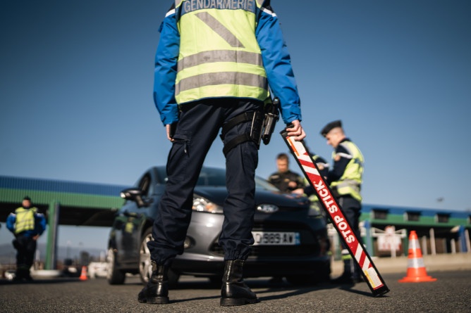 La photo représente un contrôle routier mis en place à une barrière de péage. Les gendarmes contrôlent un véhicule gris qu'ils ont arrêté.